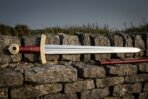 Fullered steel Viking sword with engraved brass guard and red leather grip laid along a dry stone wall above a red scabbard
