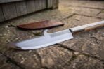 Glaive blade in close view showing the single curved edge, back hook and knotwork engraving beside a tan leather sheath