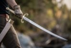 Gloved hand drawing a curved steel sabre with scrolled brass guard from a pale scabbard worn on the hip by a river