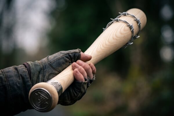 Gloved hand gripping an ash barbed wire bat by the engraved steel butt cap against a dark green forest