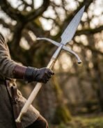 Gloved hand gripping the ash haft of a Damascus winged boar spear with ball tipped crossbar under old trees
