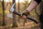 Gloved hand holding a Damascus hatchet with rune engraved head and dark wood haft in a sunlit forest
