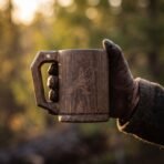 Gloved hand holding a dark hardwood beer mug with an engraved raven panel and square cut handle at sunset