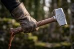 Gloved hand holding a forged hammer by its carved wooden handle, Vegvisir engraving on the steel head, mossy woods behind