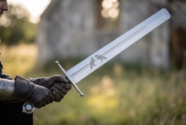 Gloved hand holding a steel arming sword with twin ravens etched on the fullered blade, ruined chapel behind