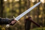 Gloved hand holding a steel medieval sword with brass guard and brass wheel pommel in a sunlit forest