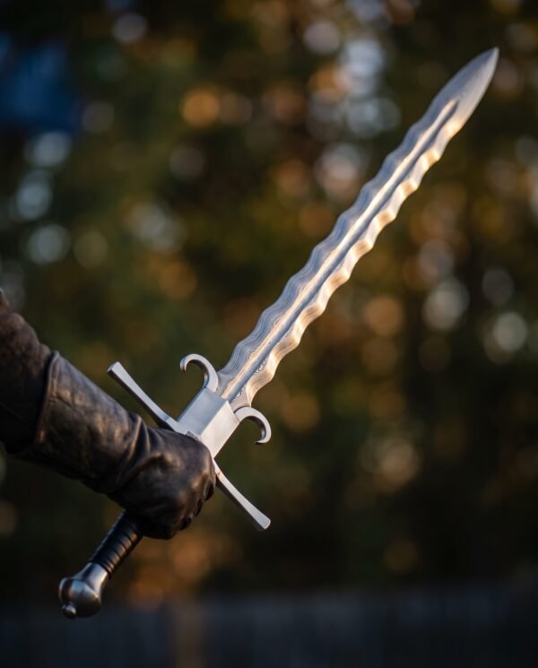 Gloved hand holding up a Damascus flamberge longsword with wavy edges and curved parrying lugs in autumn woodland