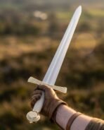 Gloved hand raising a brass hilt arming sword with wheel pommel and engraved guard against a golden hillside
