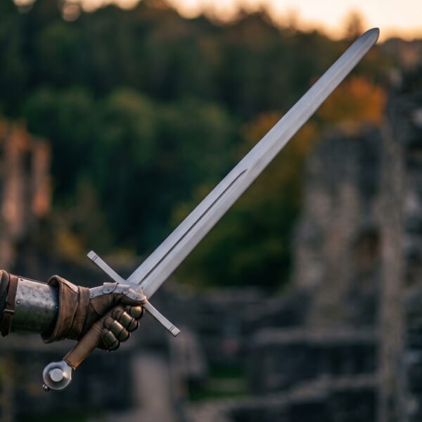 Gloved hand raising a hand and a half blunt sword with fullered blade and tan cord grip in front of ruined stone walls