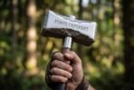 Gloved hand raising a steel war hammer with a rune engraved head against a blurred forest background