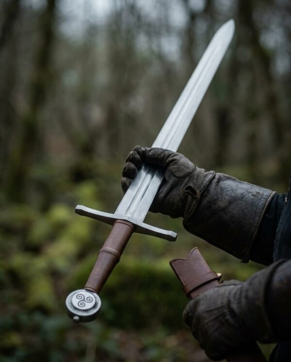 Gloved hands holding a steel arming sword with triple spiral wheel pommel and brown leather grip in a forest
