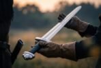 Gloved hands presenting a bright Norse sword with black grip and lobed steel pommel across a misty field