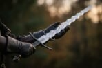 Gloved hands presenting a steel flamberge sword with wavy blade and blackened parrying hooks against a dark forest