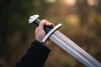 Close up of a hand gripping a Damascus sword by its black leather grip, five lobed pommel and rune marked guard in view
