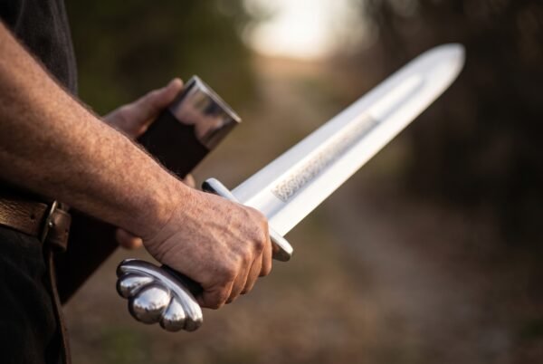 Close up of a hand gripping a Viking sword by its black leather grip, five lobed steel pommel in view, blade raised along a tree lined path