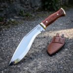 Gurkha kukri with rosewood handle and brass fittings, angled view on a gravel path with the companion knife sheath and a stone wall