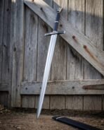 Hand and a half longsword leaning at an angle on a braced plank door with its black scabbard on the dirt below