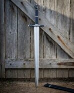 Hand and a half longsword with a faceted scent stopper pommel, a navy leather grip and a broad polished blade, leaning against a weathered timber barn door