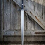 Hand and a half longsword with a faceted scent stopper pommel, a navy leather grip and a broad polished blade, leaning against a weathered timber barn door