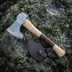 Hand axe with chevron etched steel head and pale ash haft with dark leather band lying on lichen covered rock by its edge mask