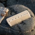 Hand carved bone viking beard comb with Celtic knotwork spine, bronze rivets and Elder Futhark runes resting on a grey boulder beside its linen drawstring pouch