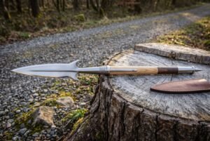 Hand forged 1095 boar spear with hooked lugs and a mirror polished head lying on a cut tree stump beside a brown leather sheath on a woodland track