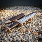 Hand forged 1095 carbon steel Roman pugio dagger with bronze guard, low angled view on gravel next to its leather sheath