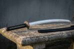 Hand forged 1095 katana sword with a polished blade showing a wavy hamon, black ito wrapped tsuka and runic compass tsuba, resting on a stone bench beside its black saya.