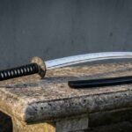 Hand forged 1095 katana sword with a polished blade showing a wavy hamon, black ito wrapped tsuka and runic compass tsuba, resting on a stone bench beside its black saya.