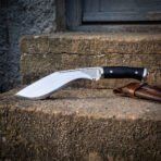 Hand forged 1095 kukri knife with black micarta handle, full length on a stone step near a doorway with its leather sheath