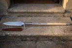 Hand forged 1095 steel boar spear with broad leaf blade and ash haft, full length on stone steps beside its leather sheath
