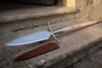 Hand forged 1095 steel boar spear with curved steel crossbar, close angled view of the blade on stone steps by its sheath