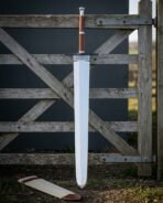 Hand forged 1095 steel broad blade arming sword with a stacked leather grip and blackened crossguard standing upright against a weathered timber field gate