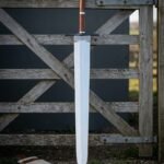 Hand forged 1095 steel broad blade arming sword with a stacked leather grip and blackened crossguard standing upright against a weathered timber field gate