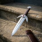 Hand forged 1095 steel fantasy dagger with winged guard and red cabochon, angled on worn stone steps beside its leather sheath