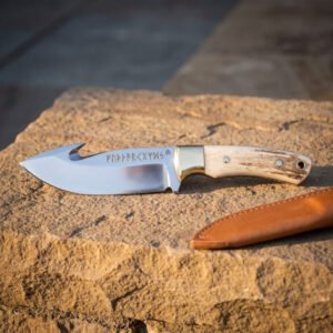 Hand forged 1095 steel gut hook hunting knife with stag antler handle, side view on sandstone beside its tan leather sheath