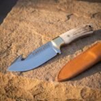 Hand forged 1095 steel gut hook knife with stag antler handle, diagonal view from above on warm sandstone with its sheath