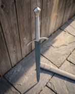 Hand forged 1095 steel knightly longsword standing point down on sunlit flagstones showing its cream cord bound grip with a triquetra pommel and arched steel crossguard
