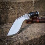 Hand forged 1095 steel kukri knife with black micarta handle, standing edge up on a stone step with the leather sheath behind