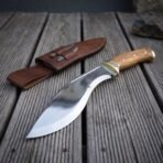 Hand forged 1095 steel kukri knife with olive wood handle and brass bolster, on wooden decking beside its leather sheath