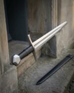 Hand forged 1095 steel medieval arming sword with faceted steel pommel and black leather grip, leaning in a stone doorway