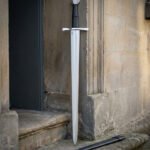 Hand forged 1095 steel medieval arming sword with fullered double edged blade, standing upright in a stone doorway beside its black scabbard