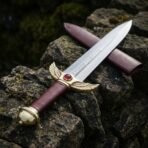 Hand forged 1095 steel rune engraved dagger with brass winged guard, angled across a mossy dry stone wall with red scabbard