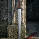 Hand forged 1095 steel serpent line Viking sword with an engraved disc pommel and russet leather grip standing against a weathered stone doorway beside its leather scabbard