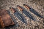 Hand forged 1095 steel throwing knives, set of three with black blades and cord grips laid diagonally on gravel by a leather roll