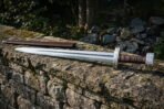 Hand forged 1095 steel viking sword with a wheel pommel, twisted brass wire and brown leather grip lying on a mossy dry stone wall in woodland