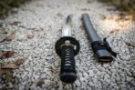 Hand forged 1095 steel wakizashi short sword with cord wrapped handle, close top down view of the hilt and saya on gravel