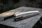 Hand forged 1095 steel wakizashi short sword with a black ribbed grip and a brown leather saya resting on a weathered garden table in evening light