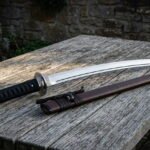 Hand forged 1095 steel wakizashi short sword with a black ribbed grip and a brown leather saya resting on a weathered garden table in evening light