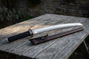 Hand forged 1095 steel wakizashi short sword with a black ribbed grip and a brown leather saya resting on a weathered garden table in evening light